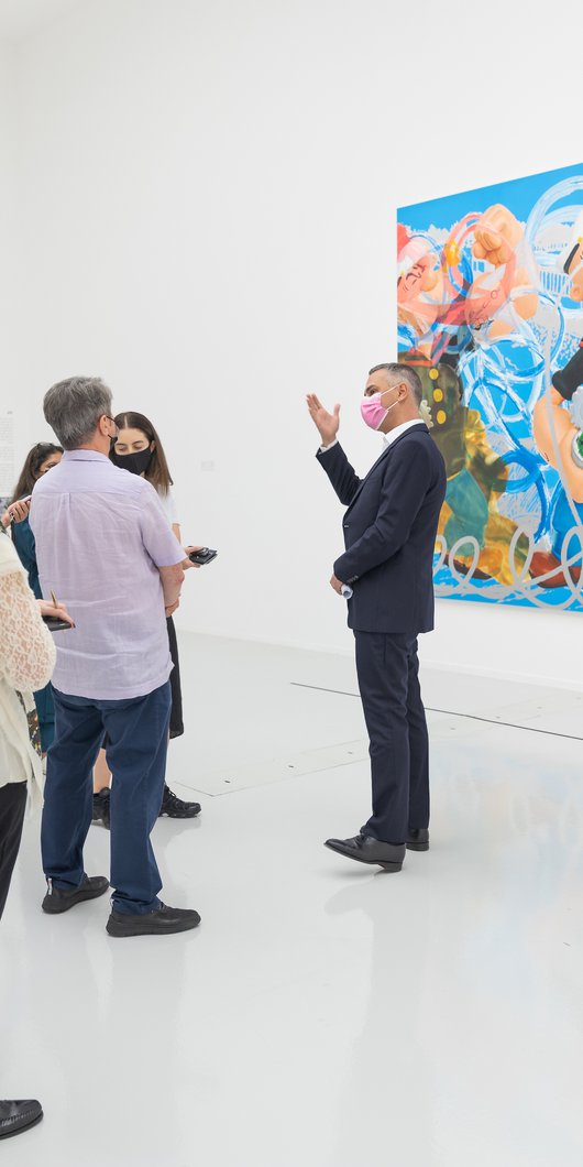 Massimiliano Gioni talking to a group of visitors in a gallery, standing in front of Jeff Koons' painting Triple Popeye