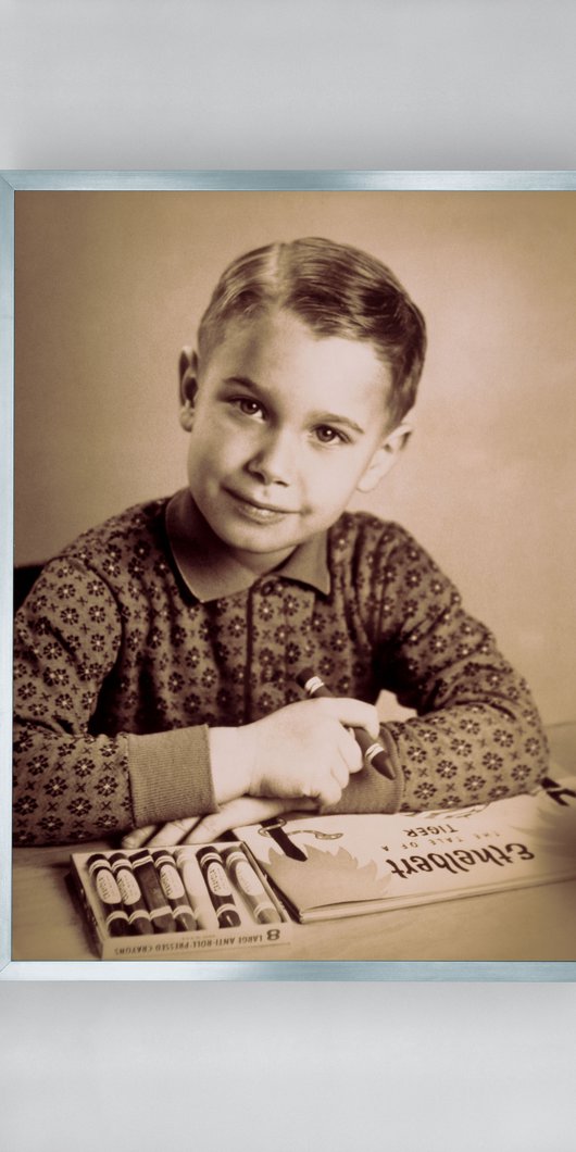 A black-and-white photograph of Jeff Koons as a child sitting at a table with a box of crayons and holding a large crayon in his right hand