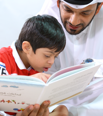 A man wearing a traditional Qatari dress reads a book to his son.