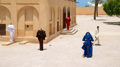 Benchellal in a white suit stands near four mannequins wearing colorful sculptural clothing outside a beige building on a sunny day.