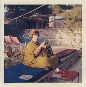 A photograph of a woman wearing a caftan sitting on a bench outside, holding her bag on her lap.