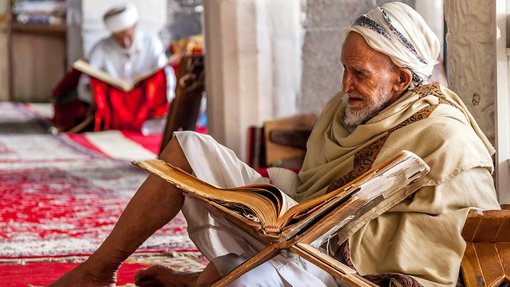 رجل مسن يرتدي الزي التقليدي يقرأ كتابًا كبيرًا على حامل خشبي في غرفة مزينة مع شخص آخر يقرأ في الخلفية.