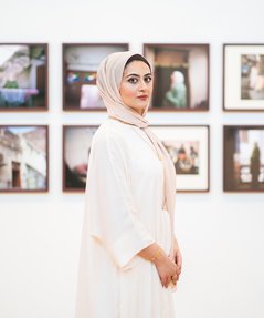 Maryam Wahid wearing white Abaya, standing in front of a wall with framed photographic works