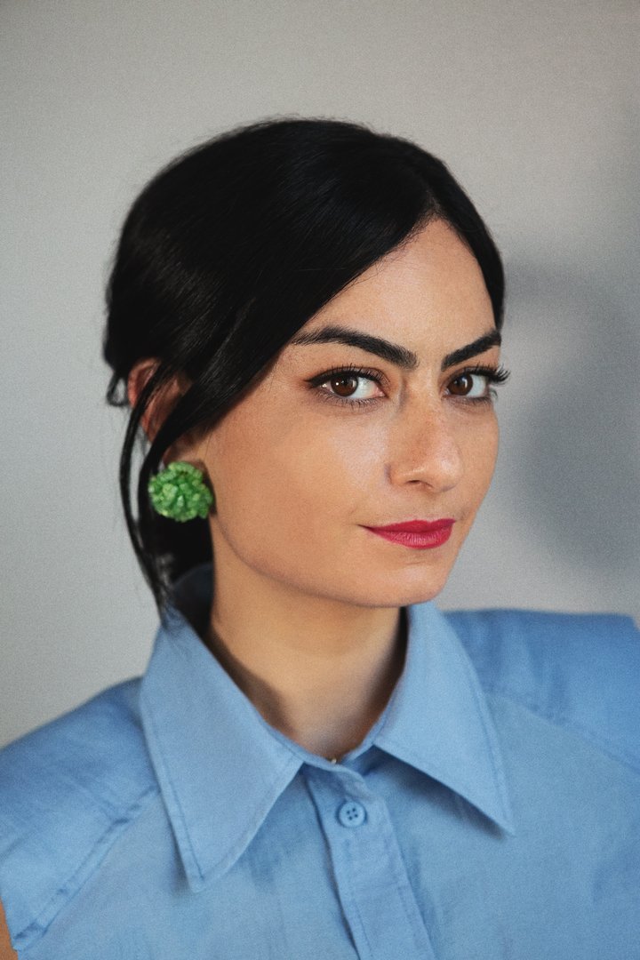 Meriem Berrada wearing blue shirt, green earrings and standing in front of grey wall