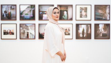 Maryam Wahid wearing white Abaya standing in front of wall with framed photographs