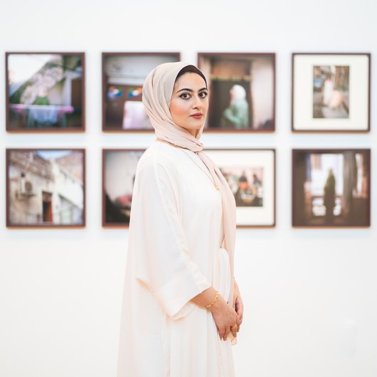 Maryam Wahid wearing white Abaya standing in front of wall with framed photographs