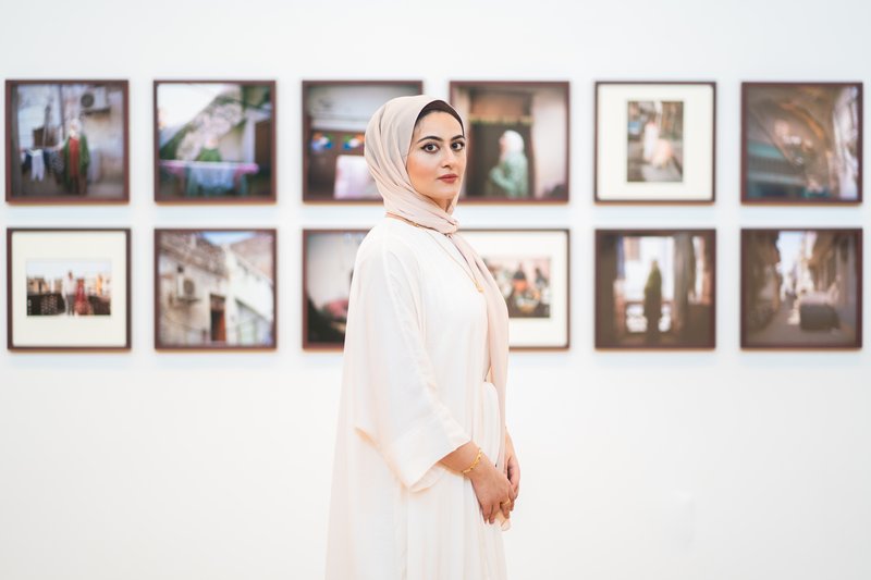 Maryam Wahid wearing white Abaya standing in front of wall with framed photographs