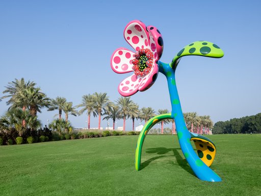 Sculpture of a big flower in multiple colours sits on a grassy hill, palm trees in the background.