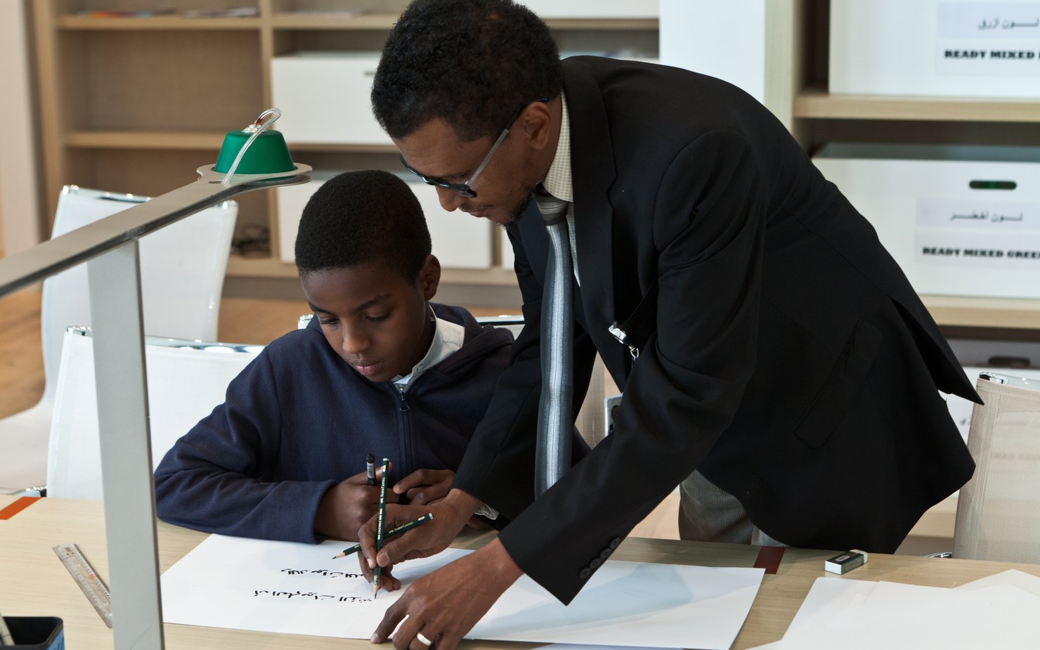 A man and child focus on drafting Arabic calligraphy letters on a piece of paper