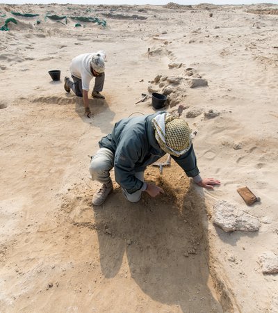 Two men kneeling on sandy desert ground using small tools to expose archeological remains at Al Zurbarah, Qatar