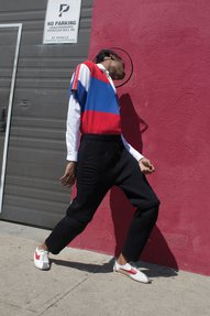 A man in a stripy top, black trousers and trainers dips and twists his body while standing next to a 'No Parking' sign.