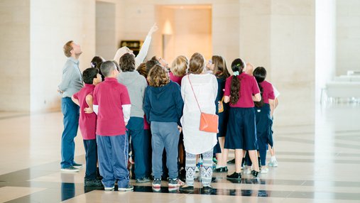 A photograph of a group of children standing in the lobby of MIA looking skywards