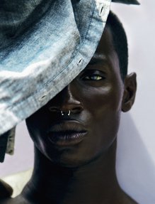 A close view of a man with a silver nose ring, looking directly at the camera, shielding his eyes with a denim shirt.