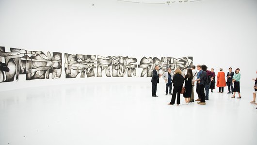 A group of visitors at the Al Riwaq's gallery space with works from Dia Al Azzawi's exhibition in the background