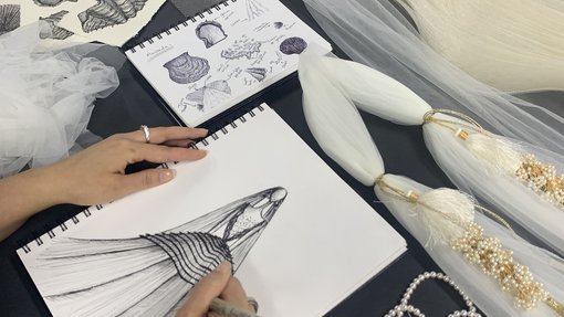 A top view of notebook with a person drawing a dress, and some fabric on a table.