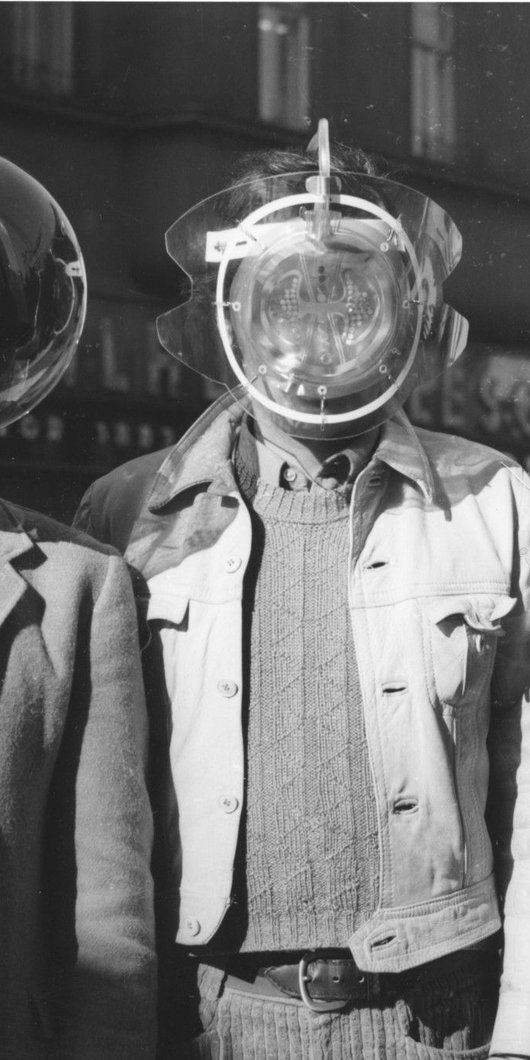 Black and white picture of three men wearing futuristic face shields and covers. Scene is from the mid- 20th century, men are wearing jackets.