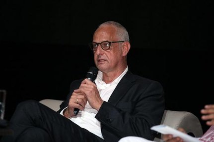 Luc Tuymans sitting on a couch while holding up a microphone to be interviewed