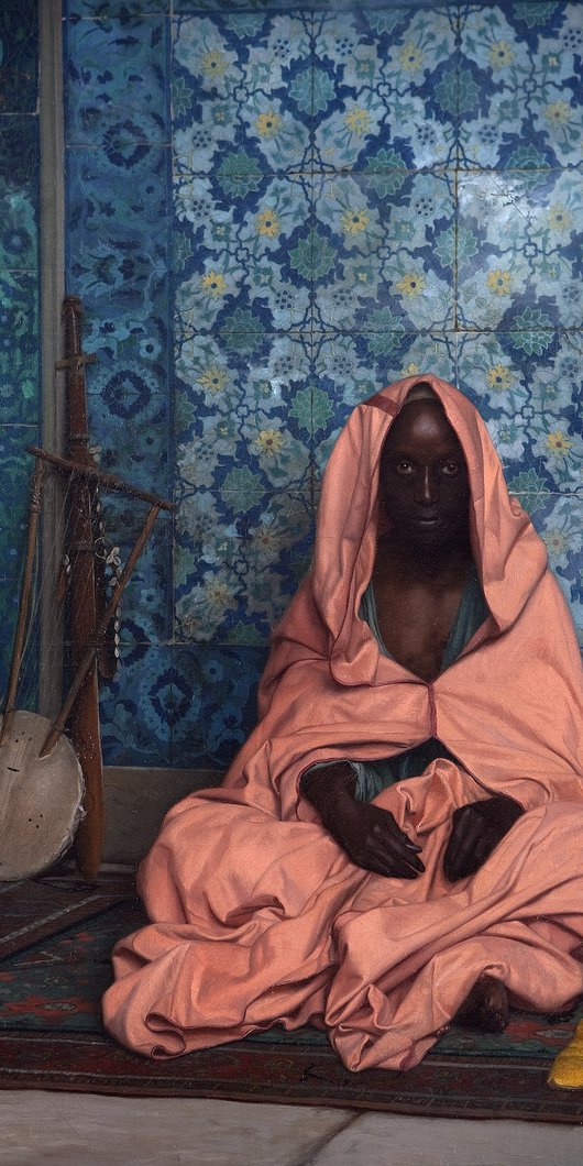 A man wrapped in pink fabric sits cross legged on a carpet, a musical instrument in the background.