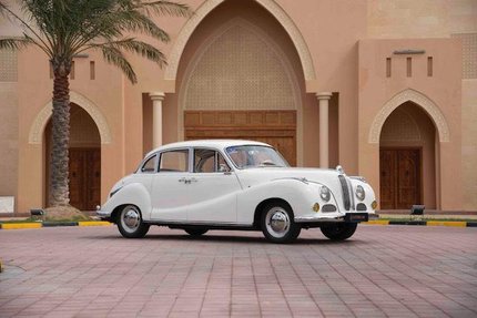 A 1955 vintage BMW car parked in front of a pale-orange building with massive, detailed arches and a palm tree on the side