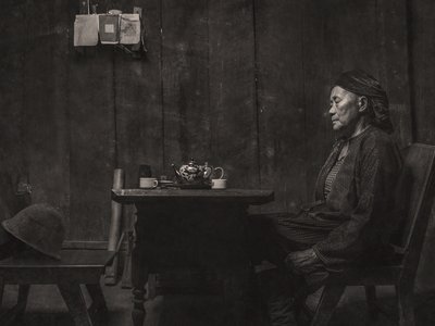 An older woman sits at a low table with a cup and teapot, facing an empty chair with a hat on the seat.