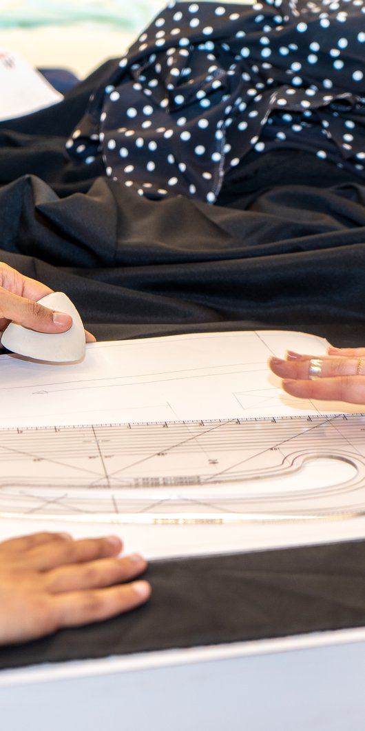 A woman drafts a pattern on top of black fabric, with another woman reviewing her work.