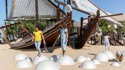 Two children in NMoQ's playground standing on top of round silver balls sunken into sand with two large wooden boats (dhows) in the background