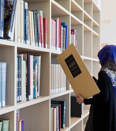 A woman holding a large thin book standing in front of white bookshelves filled with a collection of books of different colours and sizes