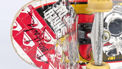 A close up of the underbelly of a skateboard, featuring red, black and yellow stickers and a black felt-tipped Tony Hawk signature.