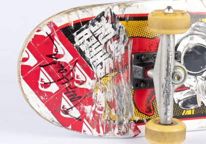 A close up of the underbelly of a skateboard, featuring red, black and yellow stickers and a black felt-tipped Tony Hawk signature.