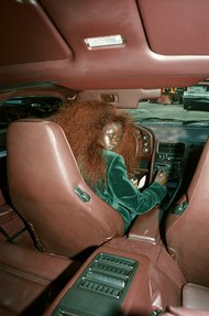 Interior view of vintage car with leather seats and a woman in the driver's seat wearing a green velvet jacket and her hair undone.