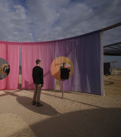 A man standing in a desert landscape looking into a round mirror with pink and purple curtains in the background